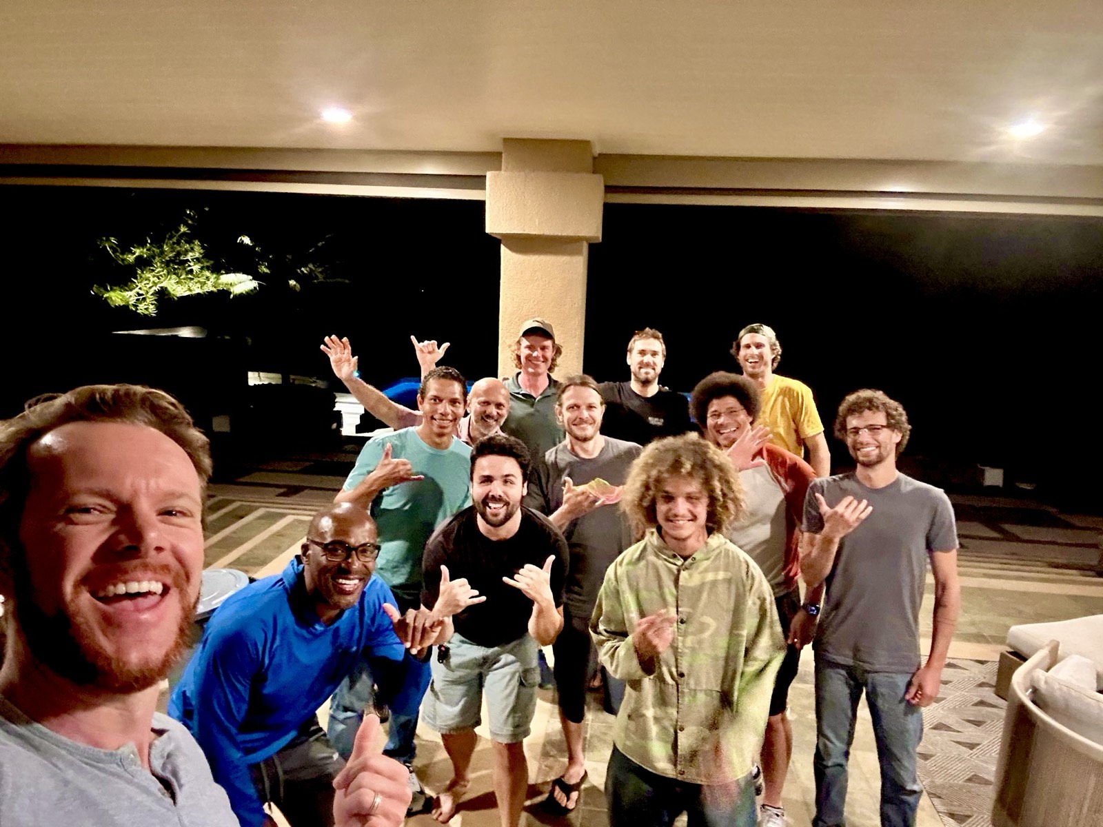A dozen men together on a covered patio at night, smiling and throwing shaka signs