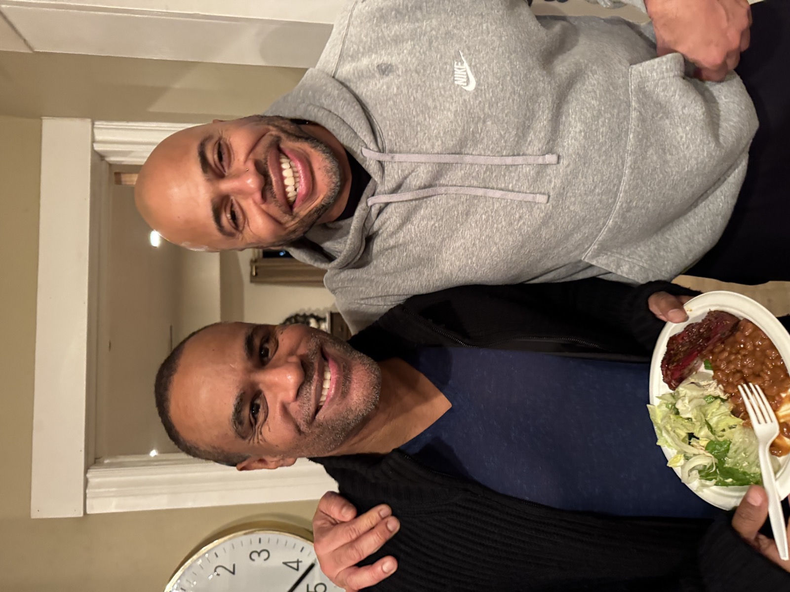 Two men sharing a meal indoors, one holding a plate, arm around the other