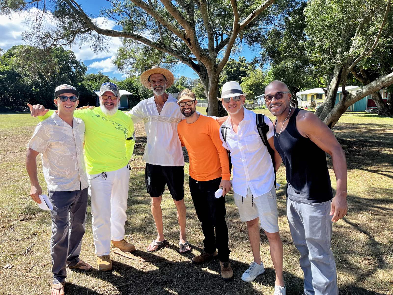 Six men under a shade tree on a grassy field, relaxed and smiling