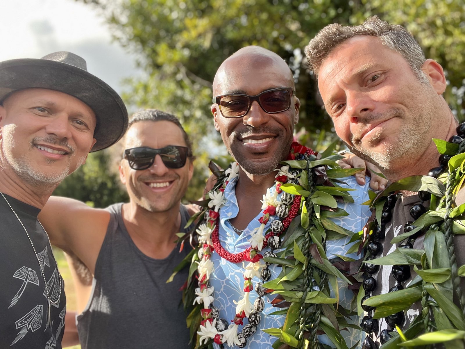 Four men in a close golden-hour photo, two wearing flower leis